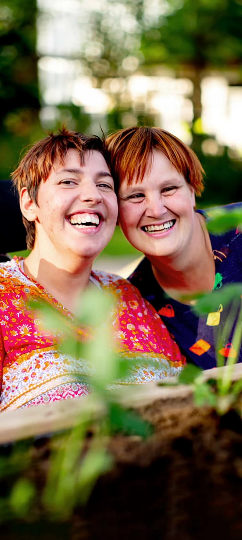 Portrait of two women in a green outdoor area. They lean their heads close together and laugh towards the camera. 